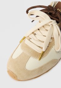 Zapatilla de ante beige y cuero crema con cordones planos crema, ojales dorados y un acento marrón texturizado. Punta redonda y suela de goma.