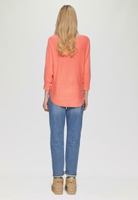 Korallfarbene Strickoberteil mit Dreiviertelärmel, kombiniert mit hellblauen geraden Jeans und beigen Knöchelstiefeln, von hinten betrachtet.