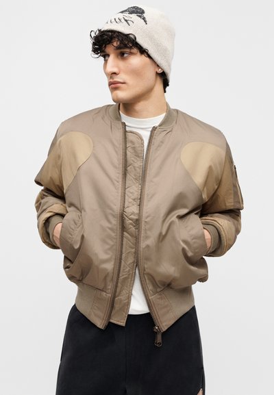 Καμηλό μπουφάν bomber με ενισχυμένες ραφές στους ώμους, εμπρόσθιο φερμουάρ, ριμπ μανσέτες και τελείωμα, ελαφρύ υλικό, σε συνδυασμό με εκρού σκουφάκι.