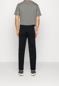 Schwarze Baumwollhose mit schmal zulaufender Passform, kombiniert mit einem grauen Kurzarm-T-Shirt. Das Modell trägt schwarz-weiße Sneakers.