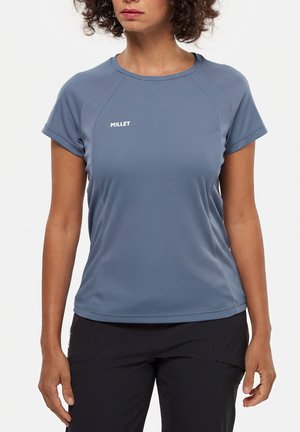 Femme portant un t-shirt de sport bleu à manches courtes de la marque Millet et un pantalon noir, debout devant un fond blanc uni.