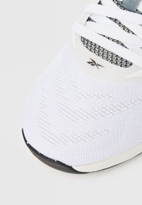 Lähikuva valkoisen urheilukengän varpaasta, jossa on musta kumipohja, teksturoitu neulekuvio, valkoiset nauhat ja pieni musta logo ylätyynyllä.