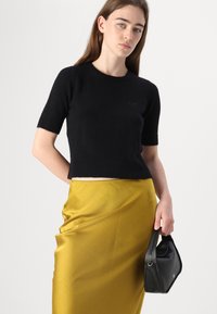 Svart kortermet strikket topp med ribbestrikkede mansjetter og nederkant, med en subtil logo. Kombinert med et skinnende gult skjørt og en liten svart veske.