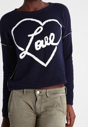 Marineblå cropped sweatshirt med hvid "Love" skrift inde i et hjerte-design. Materialet har en blød tekstur og er parret med olivengrønne bukser.