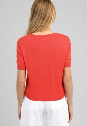 Femme aux longs cheveux blonds portant un pull rouge vif à manches courtes et un pantalon blanc, vue de dos sur un fond gris.