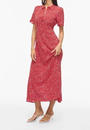 Femme portant une robe midi rouge à manches courtes avec des pois blancs et des sandales blanches à talons ouverts sur un fond uni.
