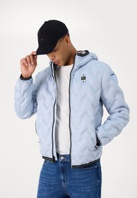 Giacca imbottita azzurro chiaro con un motivo a zig-zag, zip nera e cappuccio. Indossata con una maglietta bianca e jeans blu scuro, completata da un cappellino nero.