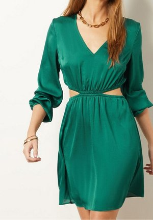 Femme portant une robe verte à manches longues avec un col en V et des découpes à la taille, accessoirisée d'un collier en chaîne dorée et de bracelets.