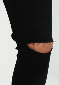 Svarta denimleggings med en sliten knädel, som visar en stor reva med fransade kanter, vilket avslöjar huden därunder.