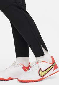 Schwarze Sporthosen mit seitlichen Reißverschlüssen, kombiniert mit weißen und orangefarbenen Nike-Schuhen, die eine Mesh-Oberfläche und gestreifte Akzente in leuchtendem Gelb aufweisen.