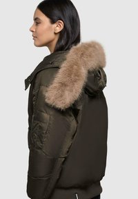 Brun puffer-jakke med en falsk pels hætteaccent. Den har en lynlåslomme og en quiltet tekstur, der viser en tætsiddende design.