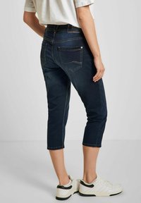 Tmavě modré denimové capri kalhoty s bílým prošíváním, vybavené zadními kapsami a pasem střední výšky. Nosené s bílými teniskami.