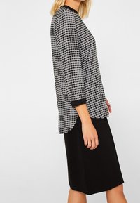Schwarz-weißer Hahnentrittmuster-Oberteil mit Dreiviertelärmel und schwarzem Kragen, kombiniert mit einem schlichten schwarzen knielangen Rock.