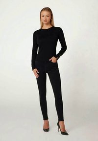 Frau steht, trägt ein eng anliegendes schwarzes langärmliges Oberteil, schwarze enge Hosen und schwarze High Heels vor einem schlichten hellen Hintergrund.