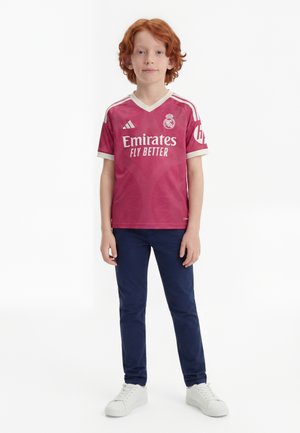 Ein rothaariges Kind steht und trägt ein rotes Real Madrid Fußballtrikot, dunkelblaue Hosen und weiße Sneakers vor einem schlichten weißen Hintergrund.