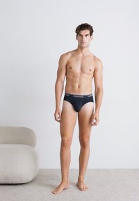 Schwarze Calvin Klein Unterhosen an einer stehenden männlichen Figur, die ein enganliegendes Design und einen weichen Bund mit dem Markenlogo präsentieren. Heller Hintergrund.