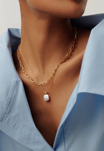 Közeli kép a nyakról, rajta réteges arany nyakláncok: egy vastag lánc és egy vékony lánc négyzet alakú fehér medállal, világoskék inget viselve.
