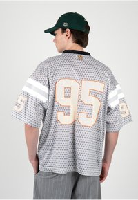 Hombre que lleva una camiseta deportiva gris con diseño y número 95 en letras grandes blancas, mangas con rayas blancas, pantalones grises de rayas finas y gorra verde oscuro.