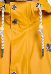 Yellow waterproof jacket featuring a high collar, drawstring with a wooden bead, silver hardware, and an emblem of a face.