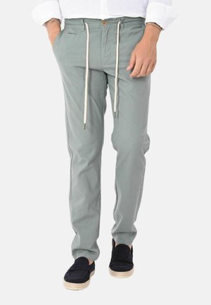 Uomo che indossa pantaloni grigio chiaro con coulisse e scarpe nere slip-on, in piedi con una mano in tasca e le maniche bianche della camicia arrotolate.