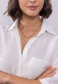 Gouden ketting met diverse kleine, kleurrijke kralen, waaronder groen en roze, gepresenteerd tegen een model dat een wit overhemd met knopen draagt.