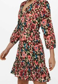 Žena nosící květované zavinovací šaty s tříčtvrtečními nafukovacími rukávy a volánkovým lemem, v růžových, zelených, modrých a béžových barvách.