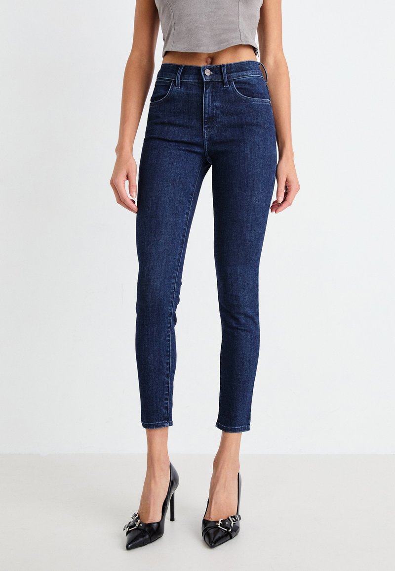 Tummansiniset skinny-farkut korkealla vyötäröllä, denim-tekstuuri, viisi taskua ja lyhennetty pituus yhdistettyinä mustiin korkokenkiin.