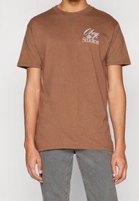 Hnědý bavlněný tričko s krátkým rukávem a kulatým výstřihem, s bílým logem "Obey Studios" na levé části hrudi.