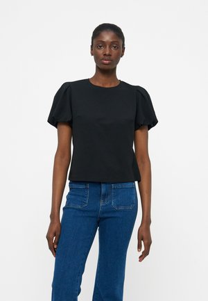 Γυναίκα που φοράει μαύρη μπλούζα με φουσκωτά μανίκια και μπλε τζιν, στέκεται μπροστά σε απλό λευκό φόντο.