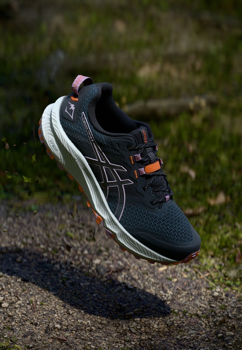 Chaussure de course bleu foncé et orange suspendue au-dessus d'un chemin de gravier avec un fond flou d'herbe, mettant en valeur le design de la chaussure et la texture de la semelle.