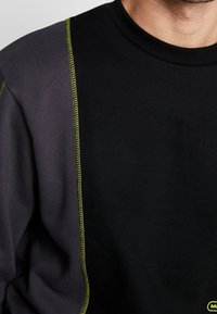 Sudadera negra y gris oscuro con costuras en contraste de color amarillo fluorescente a lo largo del hombro. Cuello acanalado. Textura suave.