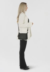 Borsa a tracolla nera in pelle testurizzata, con hardware argentato e un'etichetta staccabile. Indossata con un blazer bianco e pantaloni neri aderenti.
