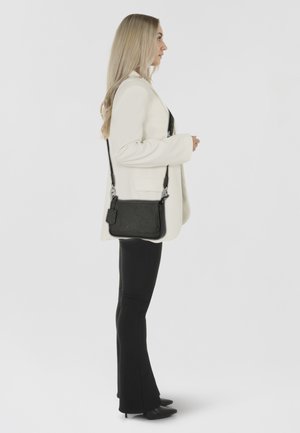 Borsa a tracolla nera in pelle testurizzata, con hardware argentato e un'etichetta staccabile. Indossata con un blazer bianco e pantaloni neri aderenti.