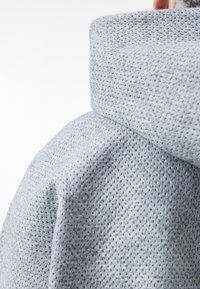 Ljusgrå, texturerad tyg med ett andningsbart nätmönster. Har en hög krage och mjuk fall, med fokus på komfort och funktionalitet.