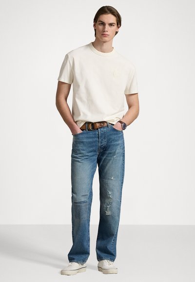 Camiseta de cuello redondo color beige claro con un logo sutil, combinada con jeans azules desgastados, un cinturón marrón y zapatillas blancas.