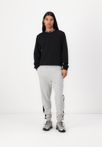 Hanorac negru cu un guler cu logo alb, combinat cu pantaloni de jogging gri cu dungi negre pe laterale și pantofi sport gri cu talpă neagră.