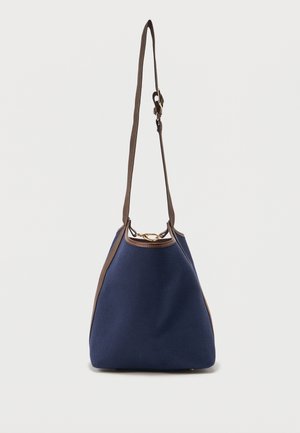 Τσάντα-κούπα σε ναυτικό μπλε με καφέ ρυθμιζόμενο λουρί ώμου, καφέ λεπτομέρειες και χρυσό κούμπωμα με δαχτυλίδι σε απλό φόντο.