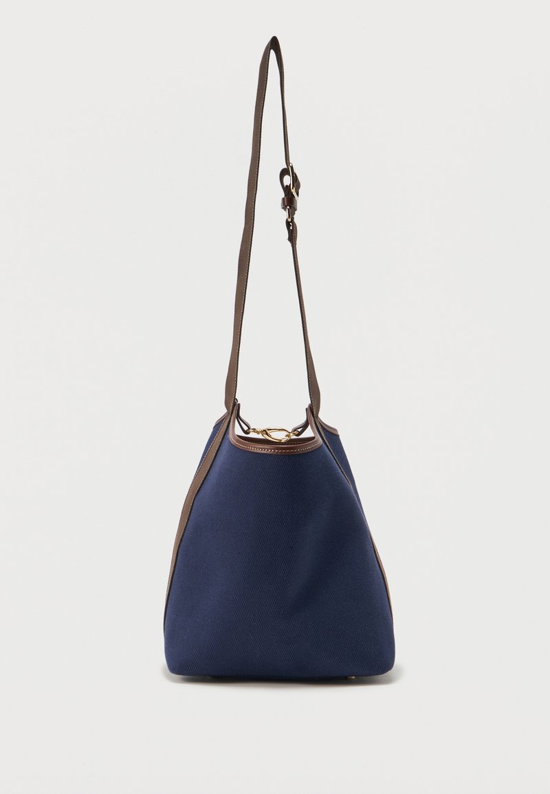 Τσάντα-κούπα σε ναυτικό μπλε με καφέ ρυθμιζόμενο λουρί ώμου, καφέ λεπτομέρειες και χρυσό κούμπωμα με δαχτυλίδι σε απλό φόντο.