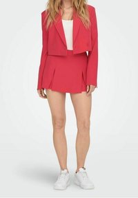 Blazer corto de color rosa brillante combinado con una minifalda plisada a juego. La tela es suave; tiene un diseño ajustado, cierre de botones y zapatos deportivos blancos.