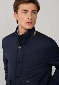 Chaqueta azul marino con cuello alto, con botones negros y un acento beige en el cuello. Incluye dos bolsillos frontales.