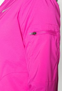 Camiseta deportiva rosa con tela texturizada, que cuenta con un bolsillo con cremallera en la parte superior del brazo y un logo de Nike en plateado cerca del hombro.