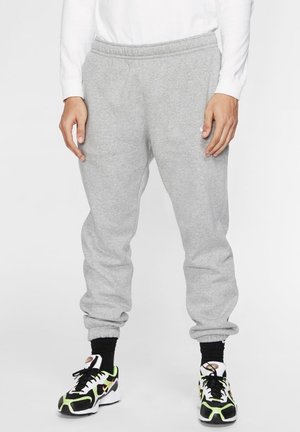 Persoon die lichtgrijze joggingbroek, zwarte sokken en witte, zwarte en limoengroene sportschoenen draagt, tegen een witte achtergrond.