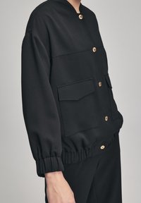 Veste noire à col en tissu lisse avec des boutons dorés, dotée de deux poches avant et de poignets élastiques pour une coupe décontractée.