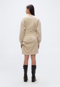 Φόρεμα φούτερ μπεζ με μακριά μανίκια και ρυθμισμένες λεπτομέρειες στη πλάτη. Συνδυασμένο με μαύρες δερμάτινες μπότες μέχρι το γόνατο.