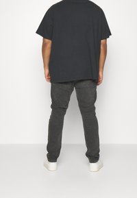 Zwarte katoenen T-shirt met korte mouwen, in combinatie met donkergrijze jeans en witte schoenen, gezien vanaf de achterkant tegen een effen achtergrond.