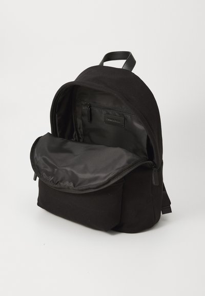Svart stoffryggsekk med en glatt, brettbar klaff. Innvendig har den en glidelåslomme og myk fôr. Enkelt design med justerbare stropper.