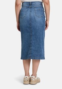 Jupe crayon en denim avec une taille ajustée et des poches arrière, dans un délavage bleu moyen, présentant un ourlet qui arrive sous le genou. Portée avec des baskets beiges.