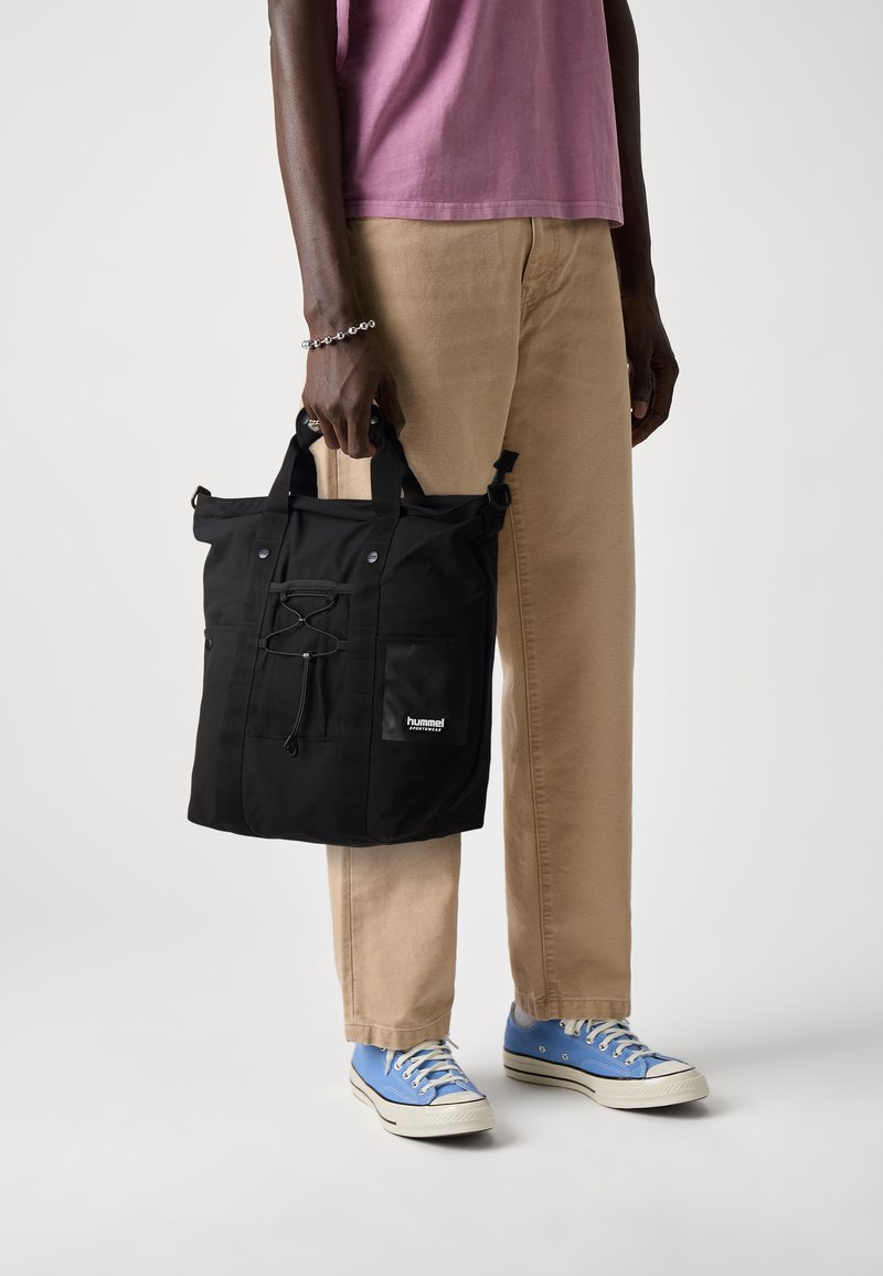 Črna torba z ročaji, ki jih je mogoče prilagoditi, sprednjim žepom in zategovalnim zaključkom. Izdelana iz trpežne tkanine, z logotipom.
