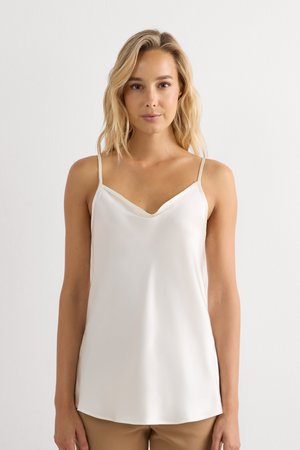 Femme aux cheveux blonds mi-longs portant un débardeur blanc sans manches et un pantalon marron clair sur un fond blanc uni.