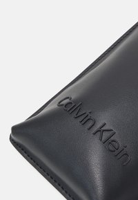 Bolsita de cuero negro con el logo "Calvin Klein" en relieve, textura suave y costuras finas a lo largo de los bordes, que se estrecha hacia un fondo puntiagudo.